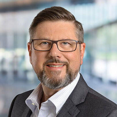 Marco Mancarella im Anzug, Hintergrund mit modernem Büroambiente, strahlt Professionalität und Freundlichkeit aus.