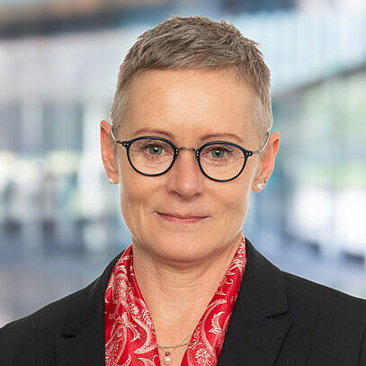 Eva Stichler mit Brille und rotem Schal, steht in modernem, lichtdurchflutetem Raum.