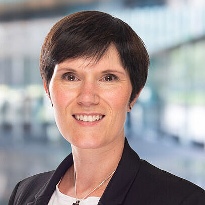 Helen Schadegg in formeller Kleidung vor verschwommenem Hintergrund. Ausstrahlung von Freundlichkeit und Professionalität.