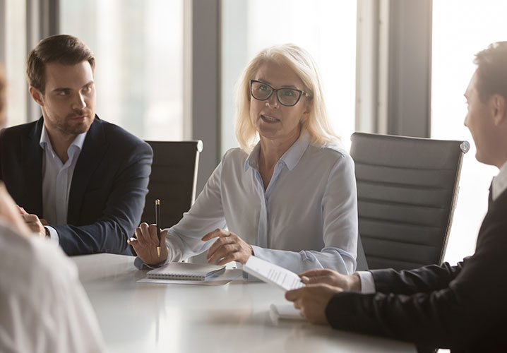 Frau spricht im Meeting, strahlt Autorität aus, Männer hören aufmerksam zu. Spannung und Konzentration im Raum.