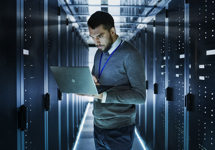 Ein Mann in einem Rechenzentrum arbeitet konzentriert an seinem Laptop zwischen Serverregalen.