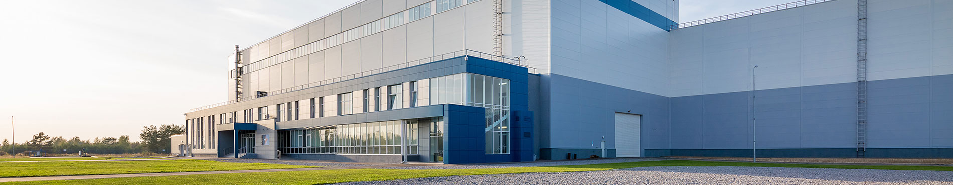 Moderne Industriegebäude mit klaren Linien, umgeben von grüner Wiese und blauem Himmel. Ruhige, sachliche Stimmung.