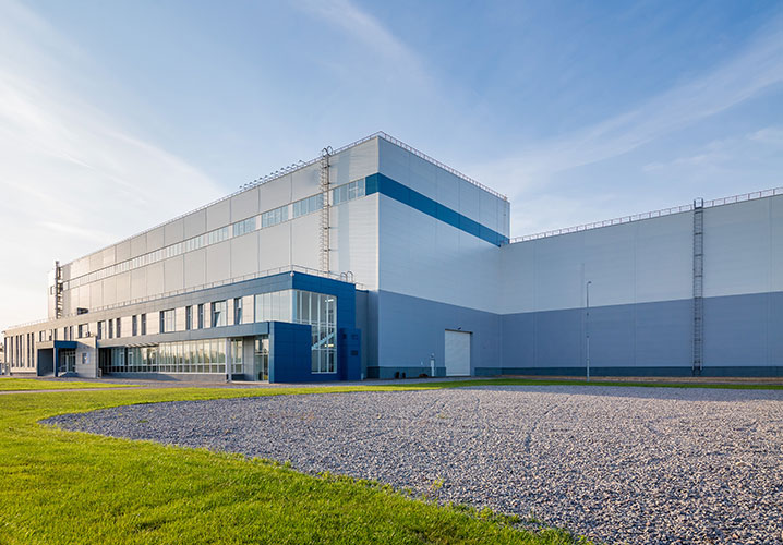Moderne Industriegebäude mit klaren Linien, umgeben von grüner Wiese und blauem Himmel. Ruhige, sachliche Stimmung.