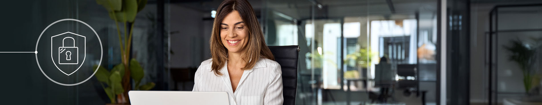 Eine lächelnde Frau arbeitet am Laptop, umgeben von Pflanzen, in einem modernen, hellen Büroraum.