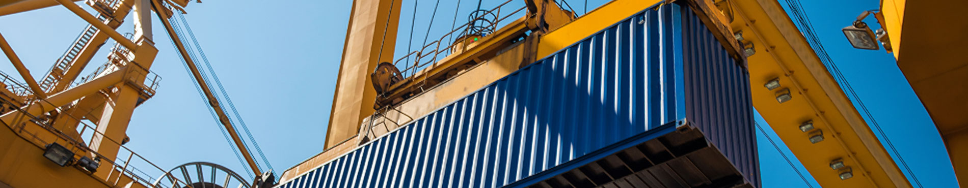 Ein blauer Container schwebt unter einem Kran, vor strahlend blauem Himmel, industrielles Ambiente.