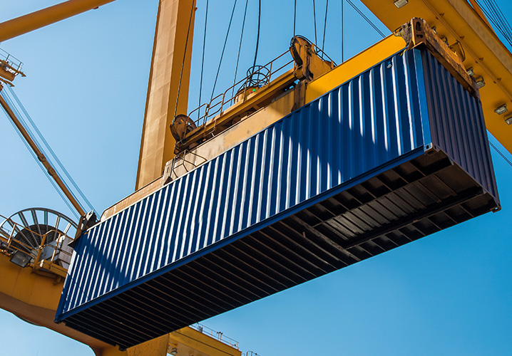 Ein blauer Container schwebt unter einem Kran, vor strahlend blauem Himmel, industrielles Ambiente.