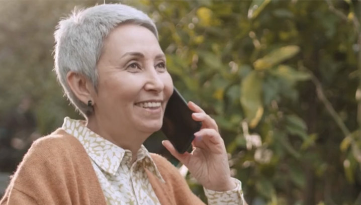 Lächelnde Frau mit grauen Haaren, telefoniert entspannt im Freien, umgeben von grüner Natur, strahlt Freude aus.