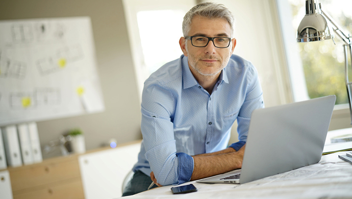 Mann mit blauen Hemd lehnt hinter seinem Laptop auf den Tisch