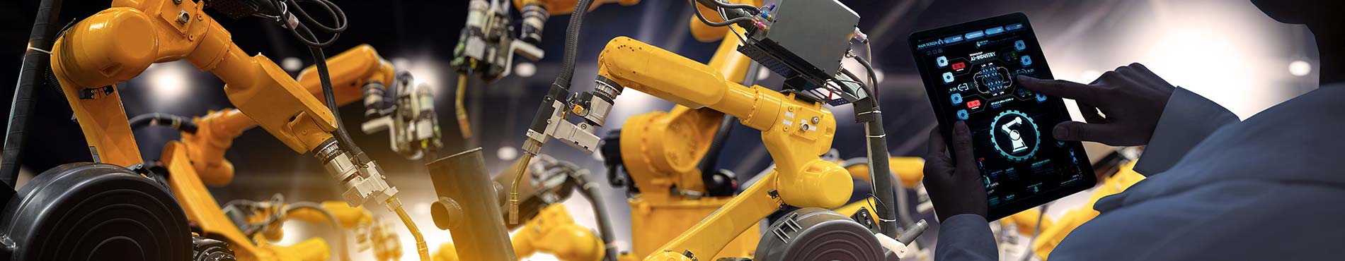 Ein Maschinenbau-Ingeneur mit einer digitalen Monitoring-Software auf einem mobilen Tablet und im Hintergrund ein automatisierter Roboterarm in einer Industriefabrik.