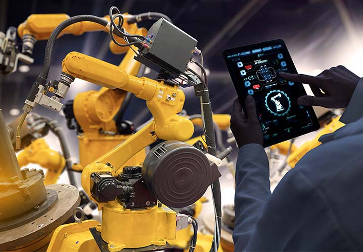Ein Maschinenbau-Ingeneur mit einer digitalen Monitoring-Software auf einem mobilen Tablet und im Hintergrund ein automatisierter Roboterarm in einer Industriefabrik.