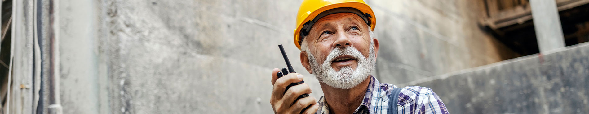 Ein älterer Bauarbeiter mit einem Funkgerät, mit einem gelben Helm in einer Baustelle.