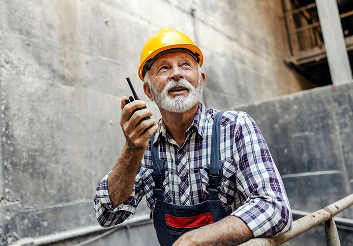 Ein älterer Bauarbeiter mit einem Funkgerät, mit einem gelben Helm in einer Baustelle.