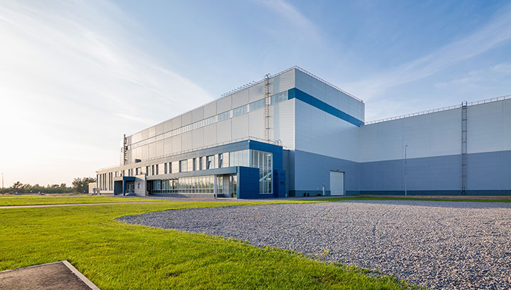 Moderne Industriegebäude mit klaren Linien, umgeben von grüner Wiese und blauem Himmel. Ruhige, sachliche Stimmung.